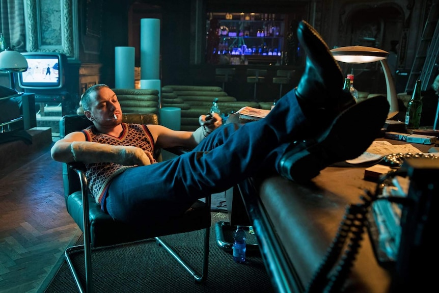 Still image of James McAvoy sitting in dark room in a chair with his feet up on a desk.