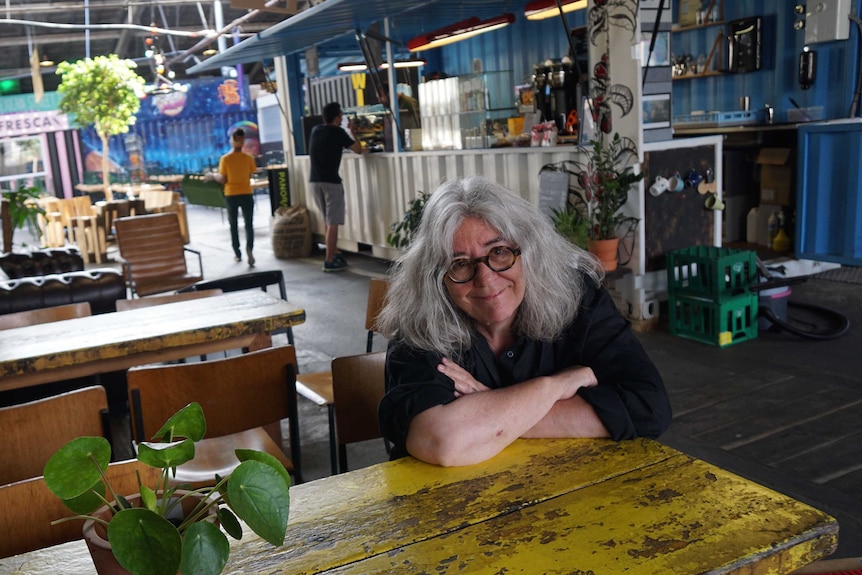 Engberg smiles at the camera with shoulder length silver hair and round lensed tortoise shell glasses, sitting in a festival bar