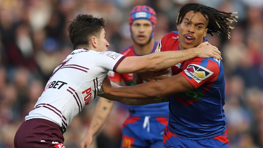 A Newcastle Knights NRL player holds the ball as he fends off a Manly opponent.