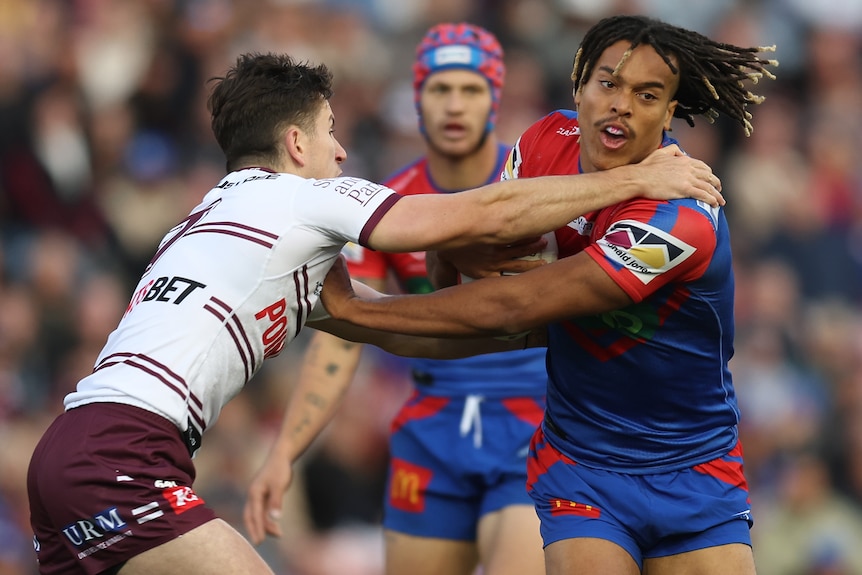 A Newcastle Knights NRL player holds the ball as he fends off a Manly opponent.