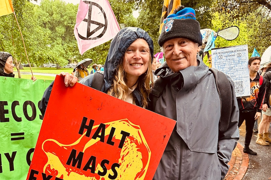 Rai and Katerina Gaita stand side by side smiling to the camera. Katerina holds a red and yellow ‘Halt Mass Extinction’ sign.