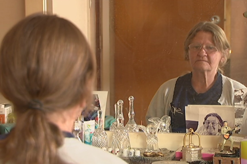 A woman sits at her dressing table and looks into a mirror.