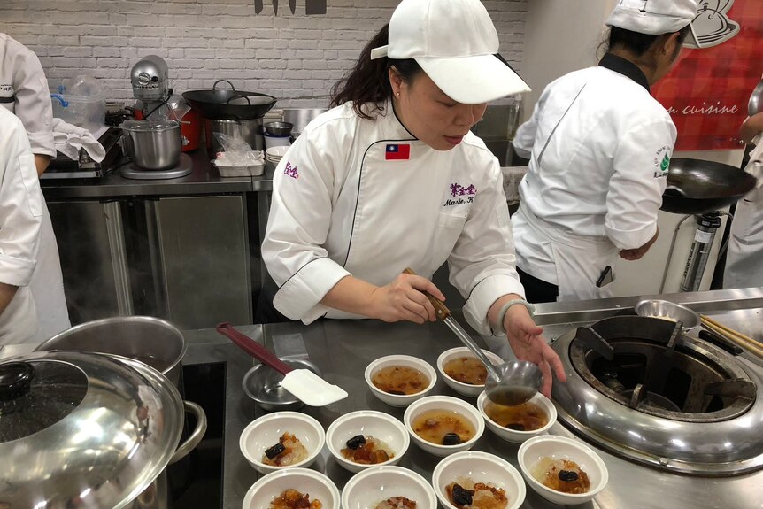 A chef ladles soup into a bowl in a commercial kitchen as other chefs cook in the background