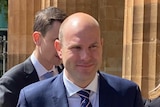 A bald man in suit and tie outside a court surrounded by reporters