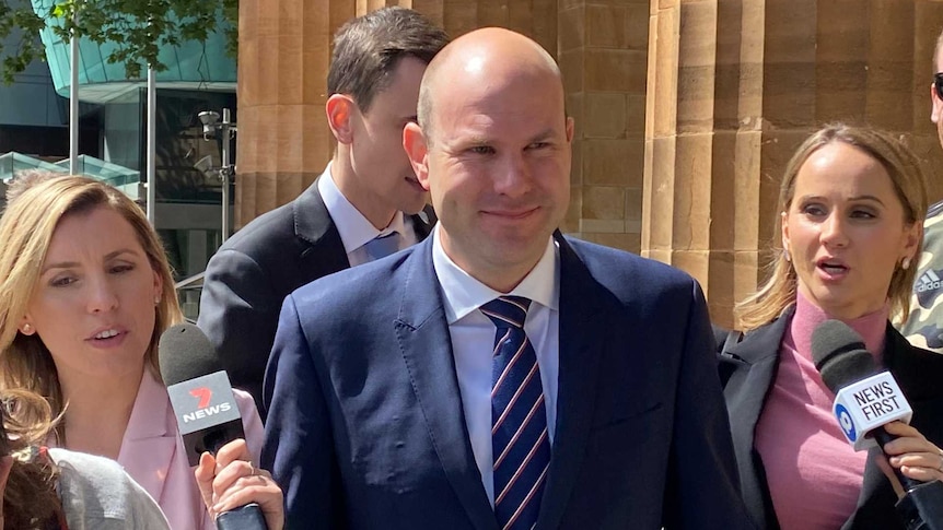 A bald man in suit and tie outside a court surrounded by reporters