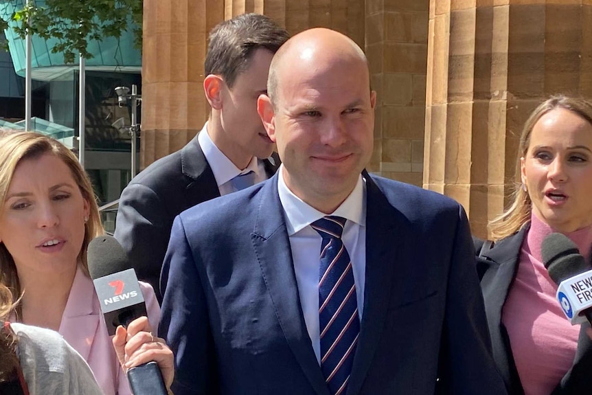 A bald man in suit and tie outside a court surrounded by reporters