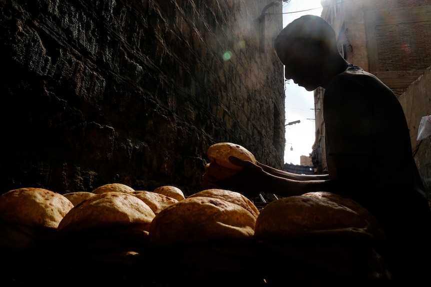 A man is seen in silhouette working in a dark alley stacking loaves of flatbread.
