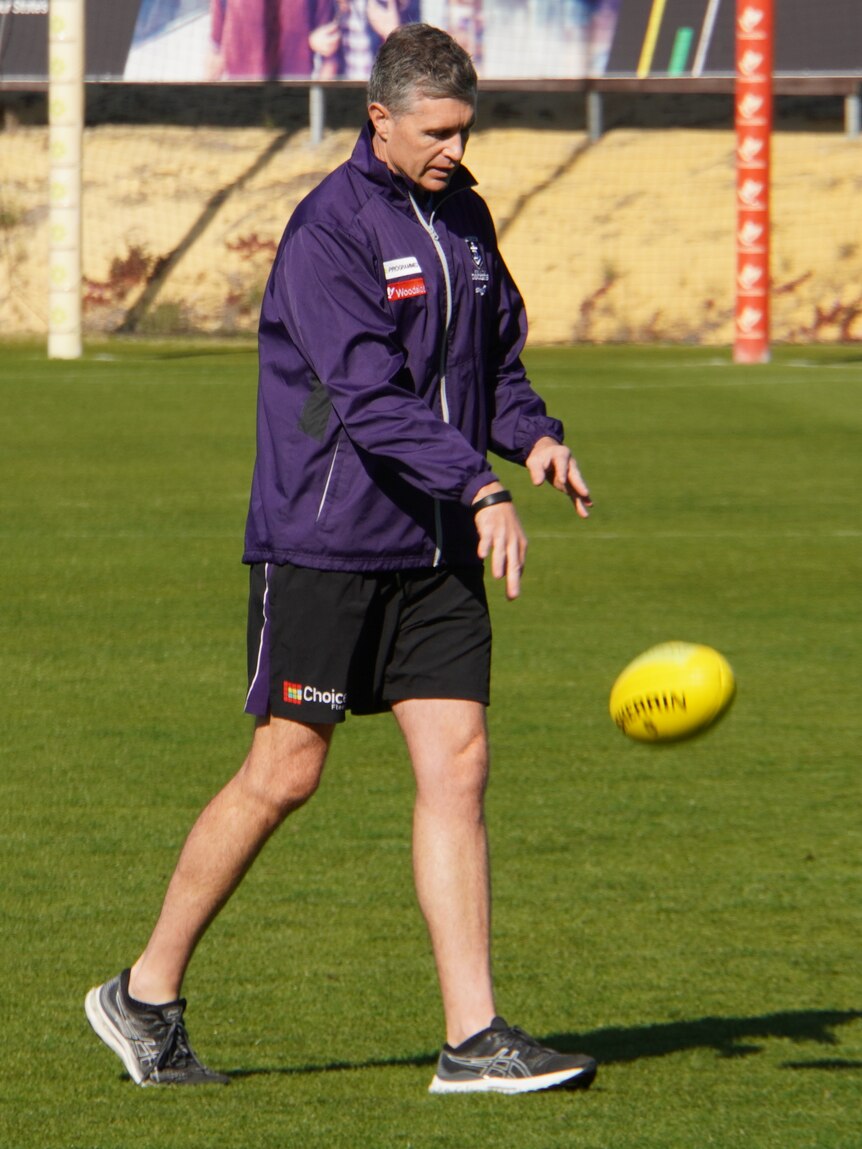 A photo of Dockers coach Justin Longmuir wearing a purple tracksuit and shorts bouncing a football at training.
