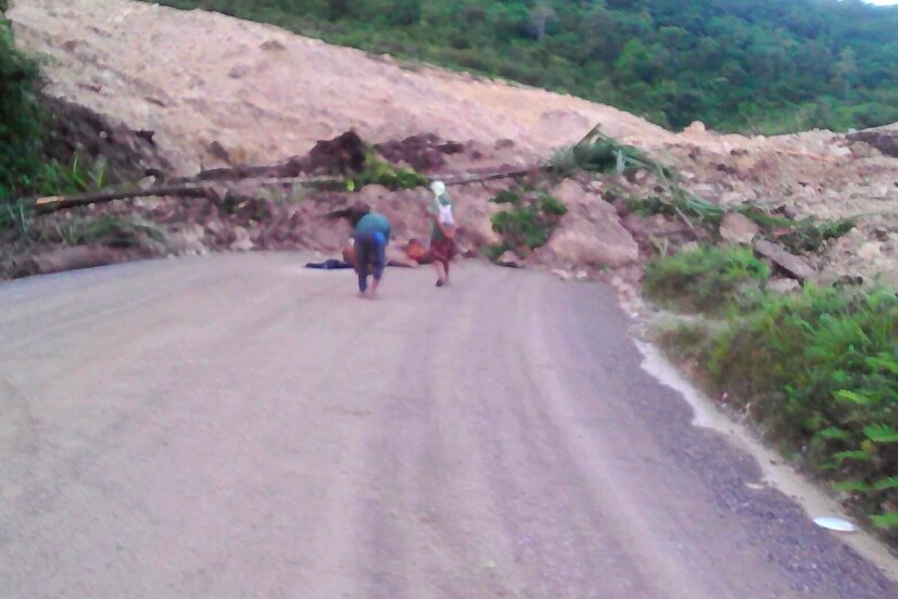 Two men stand in front of a road blocked off by a landslide.