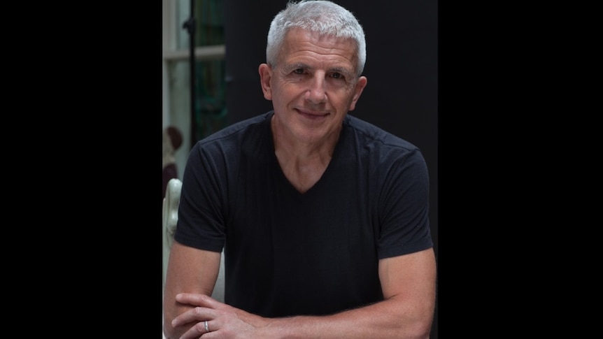 A man with short silver hair in a black t-shirt looks warmly into the camera.