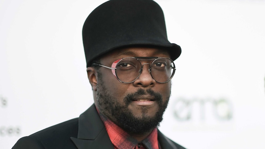will.i.am looks at the camera at an awards night wearing a black hat, suit and red button-up shirt