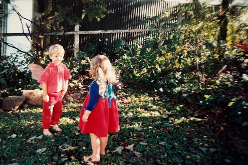 Two small children stand facing each other wearing red and blue, with matching fairy wings, in a lush subtropical yard