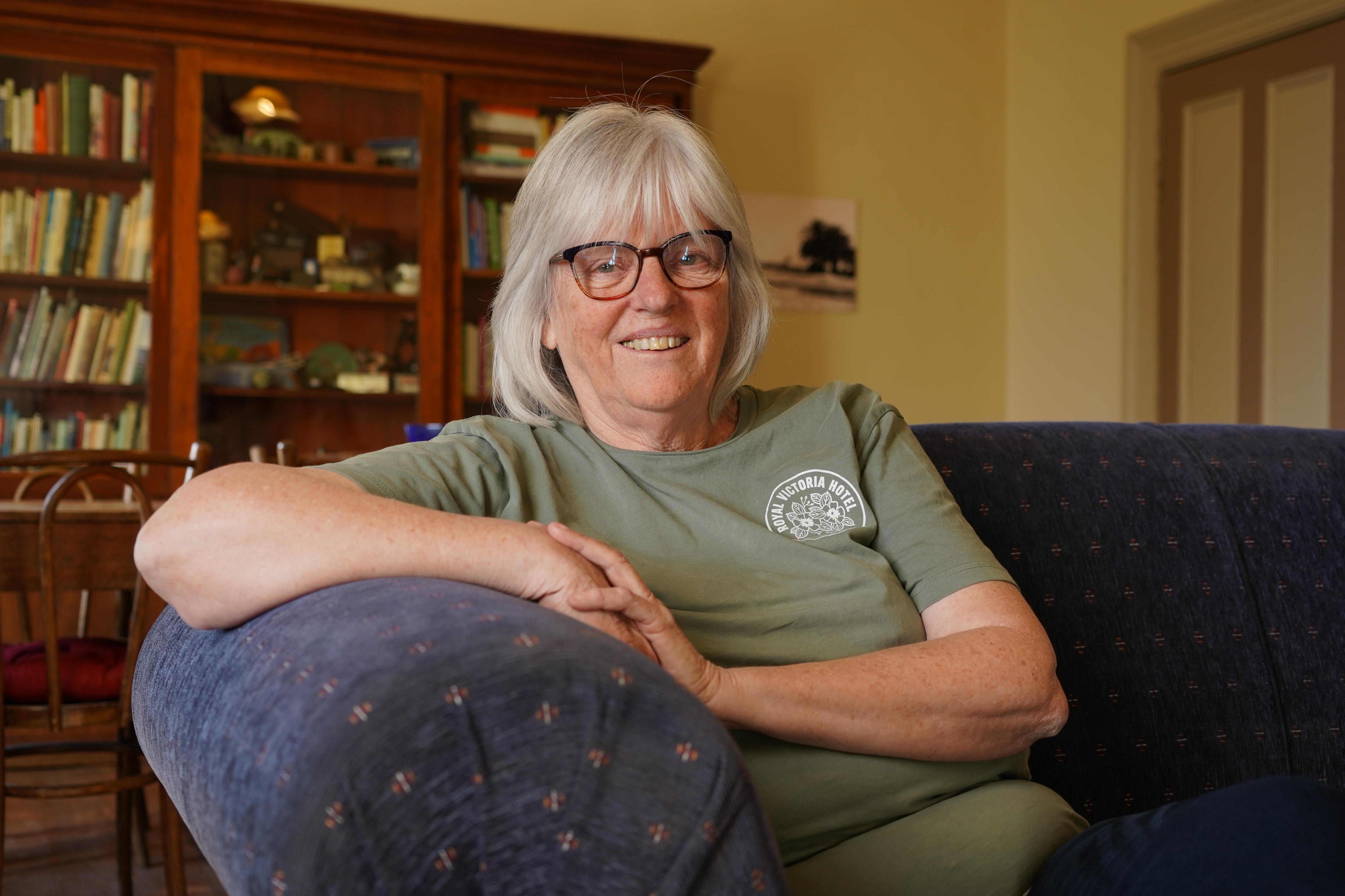A woman wearing glasses smiling sits on a blue couch inside a room with a bookcase in the background. 