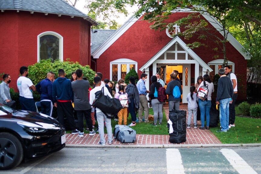 about two dozen men, women and children huddled with backpacks in front of a red church