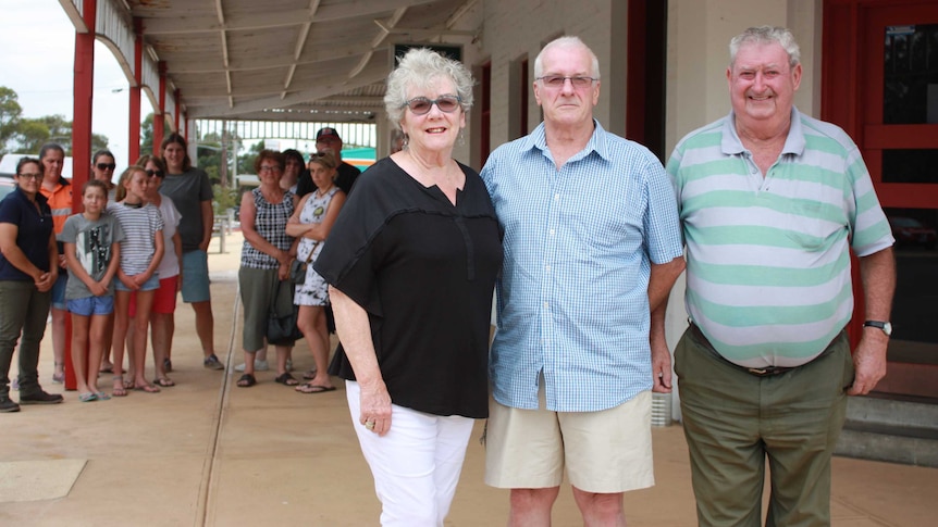 Three people stand in front of a crowd under the verandah of the Heyfield Railway Hotel.