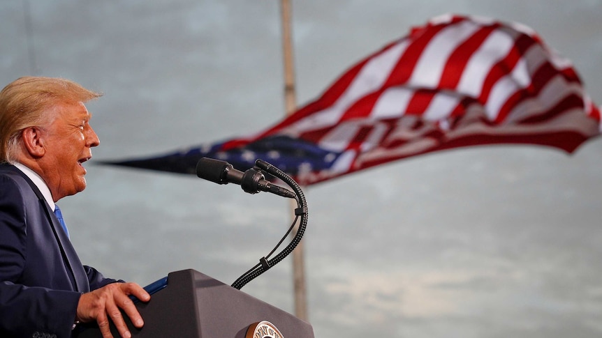 A man with orange hair speaks at a microphone while a US flag looks like it's floating out of his mouth.