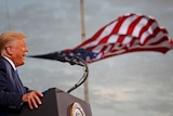 A man with orange hair speaks at a microphone while a US flag looks like it's floating out of his mouth.