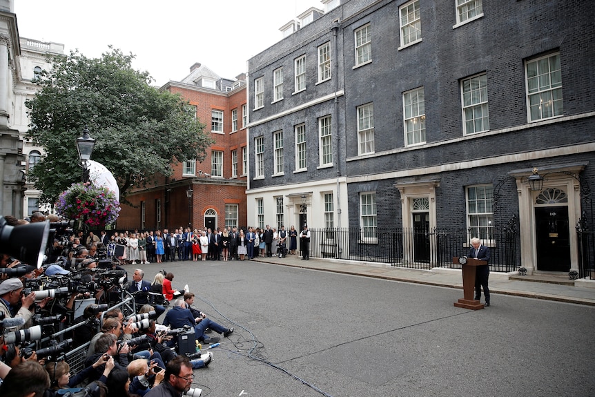 Media and Downing Street staff listen as Boris Johnson speaks at a lectern.