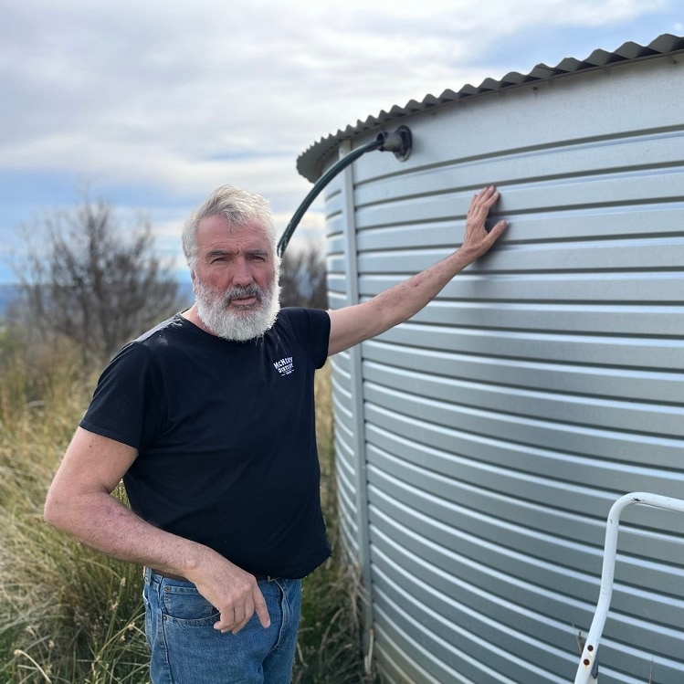 A bearded man in a black tee short standing beside a water tank on a Tasman Peninsula property
