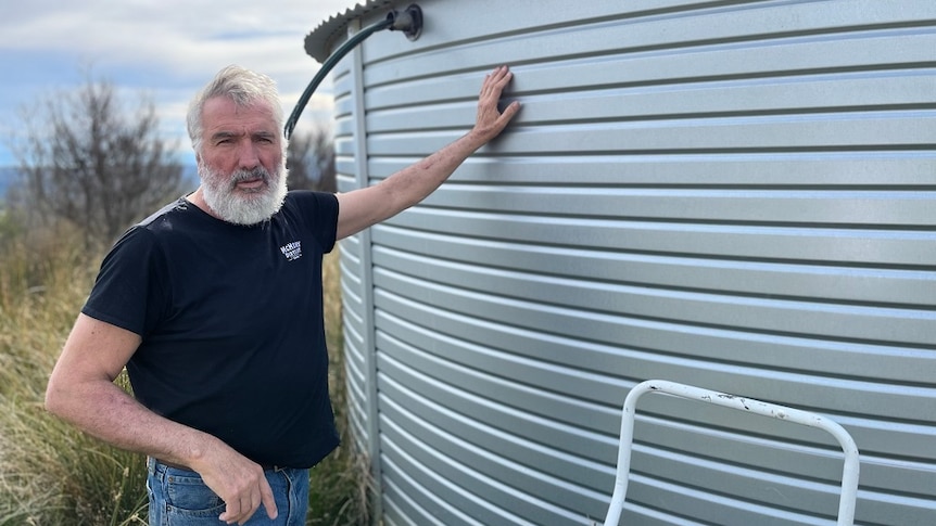 A bearded man in a black tee short standing beside a water tank on a Tasman Peninsula property