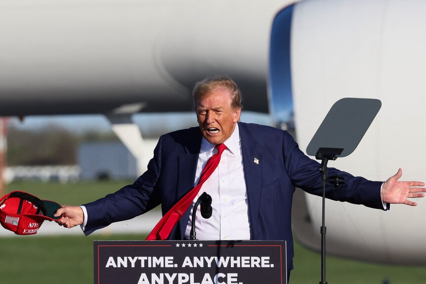 Donald Trump stands at an outdoor lectern and holds his arms out. He is holding a red cap. His red tie is flapping in the wind.