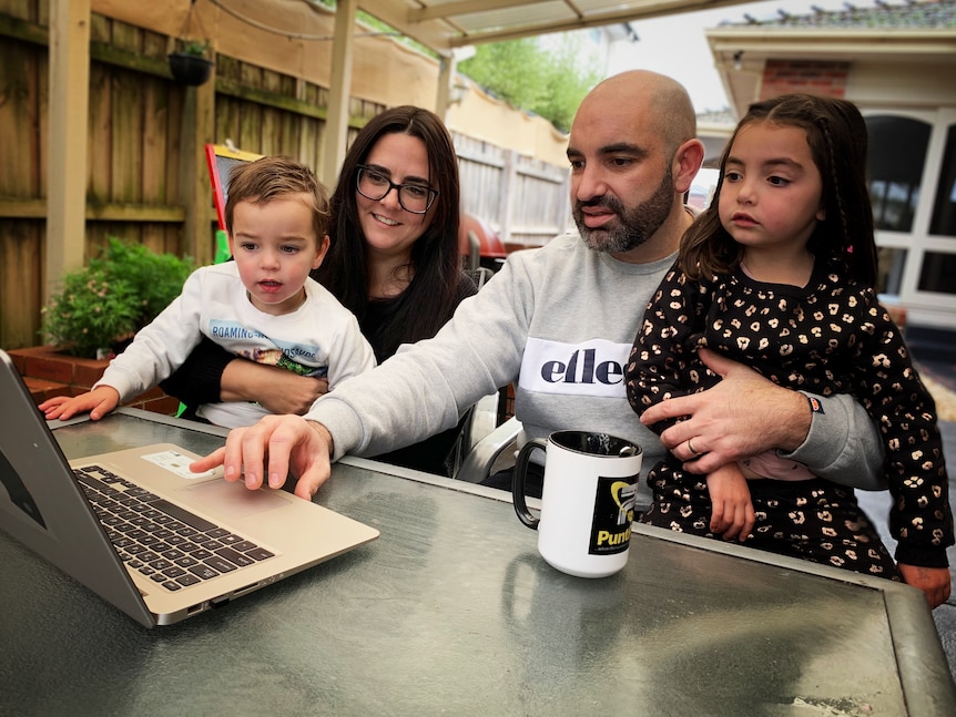 A mother and father sit at table with their two young children, looking at a laptop.