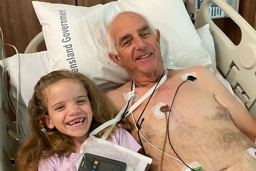 A man with grey hair in a hospital bed with wiring on his chest smiling, with a little girl next to him