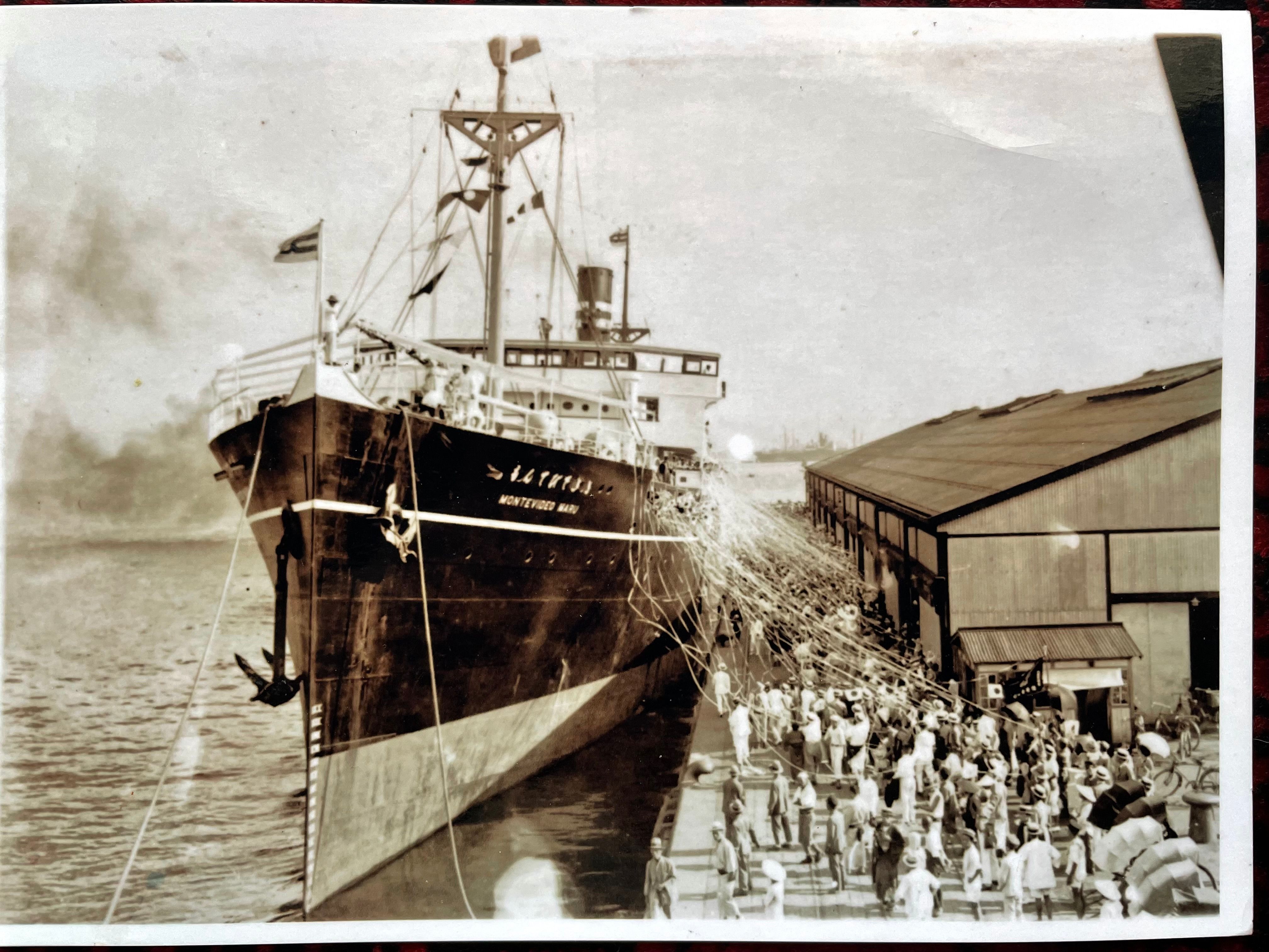 A black and white photo of a ship taking off from a harbour as people on the dock wave. 