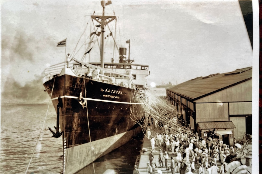 A black and white photo of a ship taking off from a harbour as people on the dock wave. 