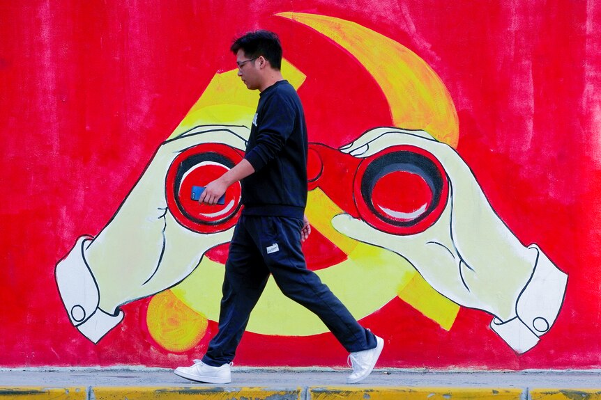 A young Chinese man in Nikes walks past a Chinese Communist Party mural with hands holding binoculars over the logo