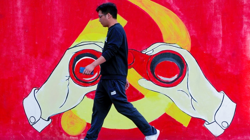 A young Chinese man in Nikes walks past a Chinese Communist Party mural with hands holding binoculars over the logo