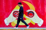 A young Chinese man in Nikes walks past a Chinese Communist Party mural with hands holding binoculars over the logo