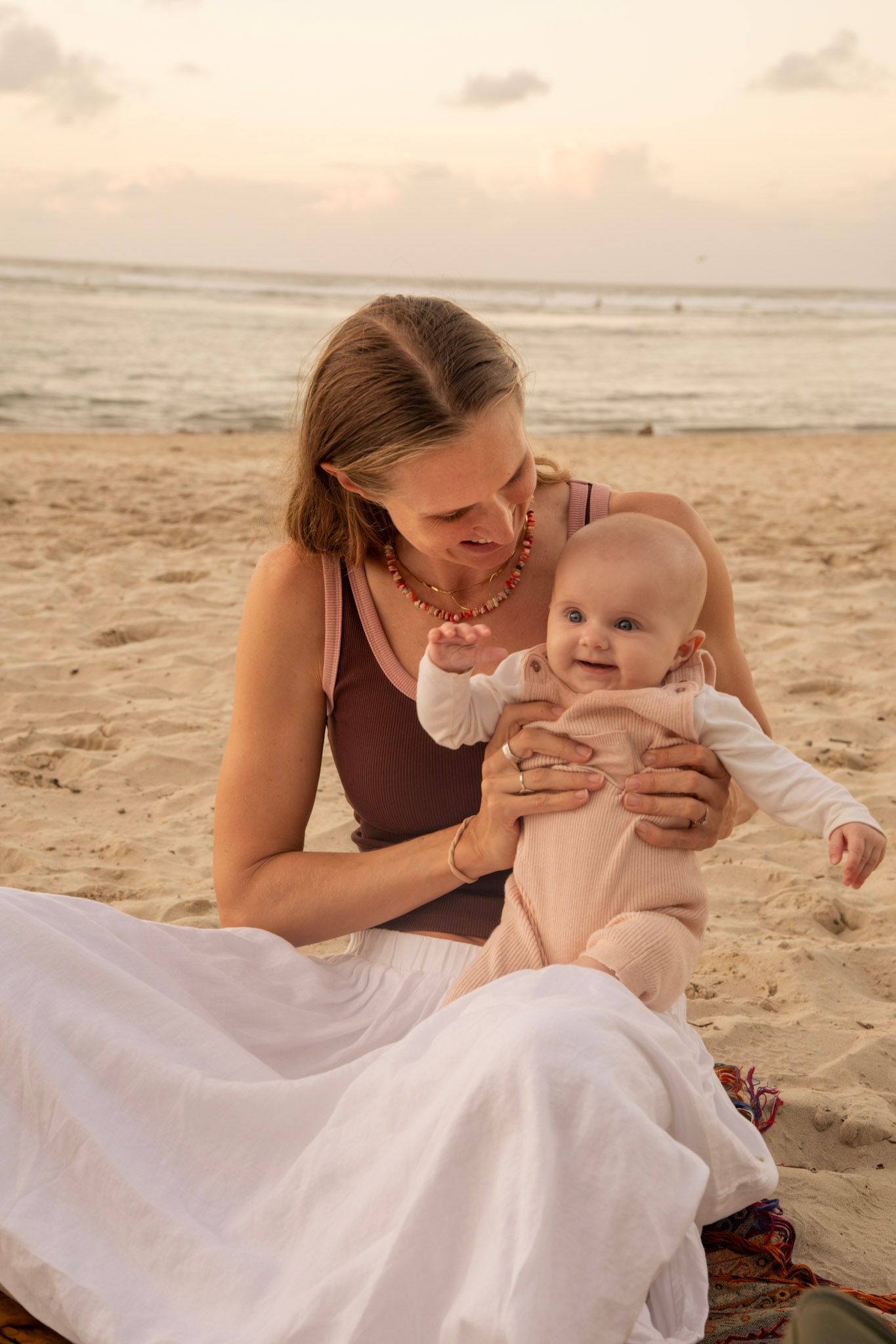 A woman looks at her baby and smiles while sitting on a beach.