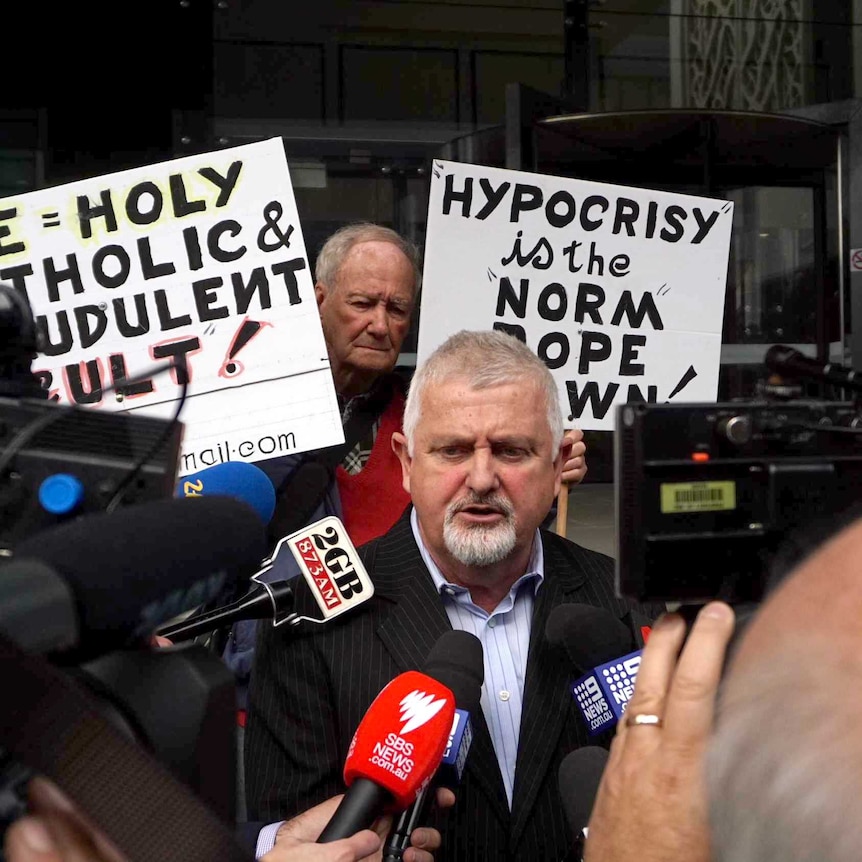 Abuse survivor Peter Gogarty speaks to the media, while supporters hold placards in the background.