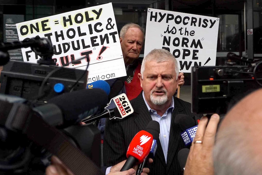 Abuse survivor Peter Gogarty speaks to the media, while supporters hold placards in the background.