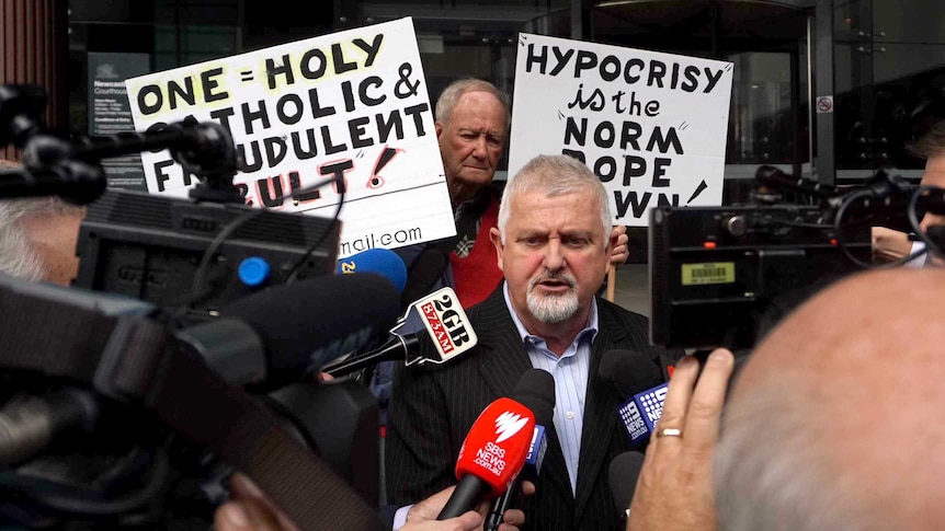 Abuse survivor Peter Gogarty speaks to the media, while supporters hold placards in the background.
