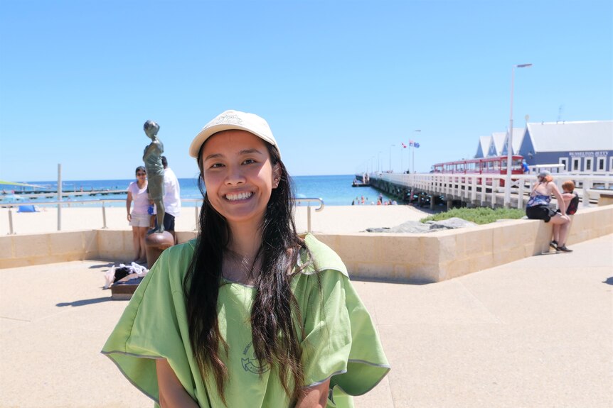 Young woman wearing cap smiles brightly in front of Busselton Jetty, clear blue skies and ocean.