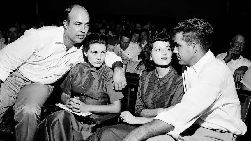 A black and white photo of two men and women, couples, sit in court looking towards one another 