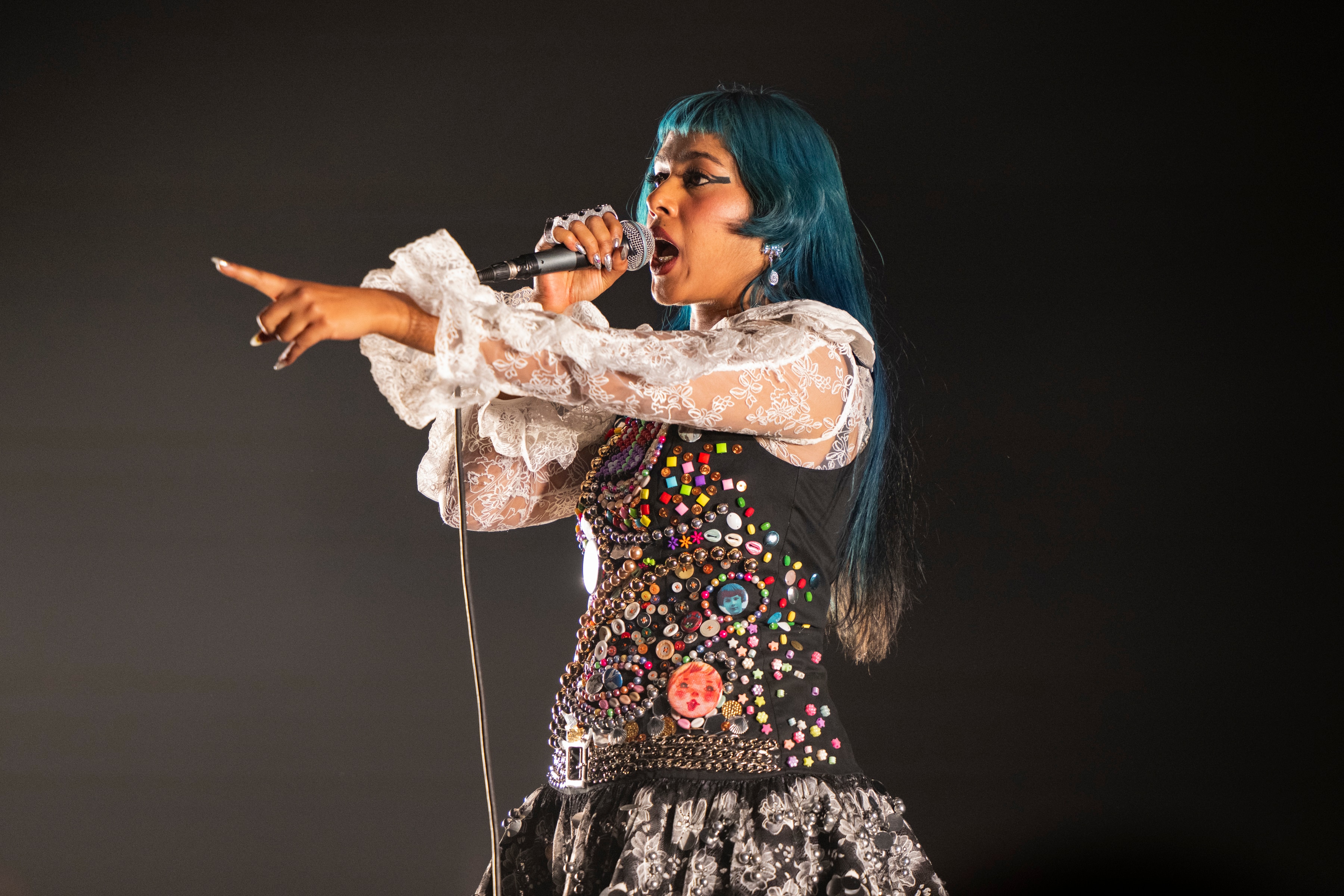 Ecca Vandal sings into a microphone on stage in a black dress with white sleeves, her blue hair is out
