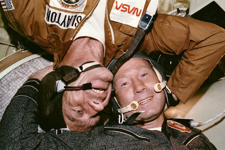 A colour photo of two men in flight helmets lying head-to-head.