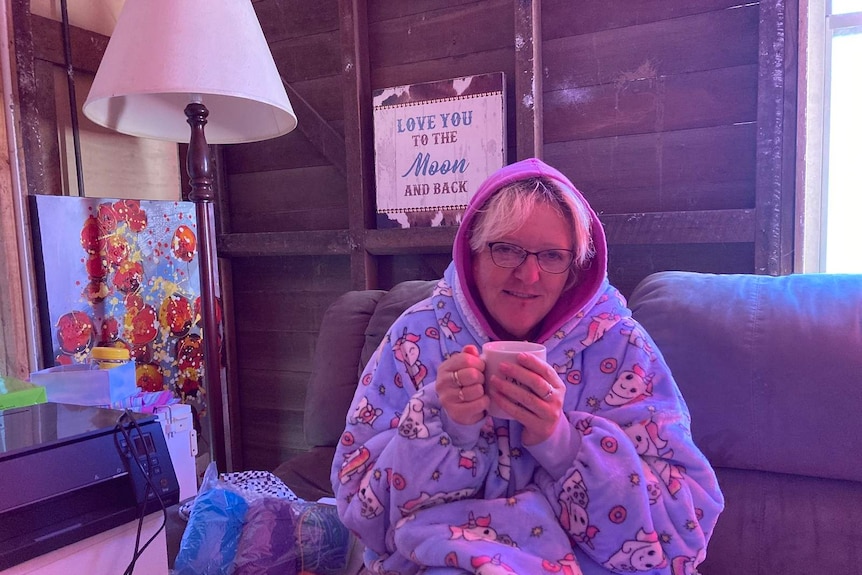A woman wears a blanket with a hood, sits on a sofa, holds a cup of coffee with both hands