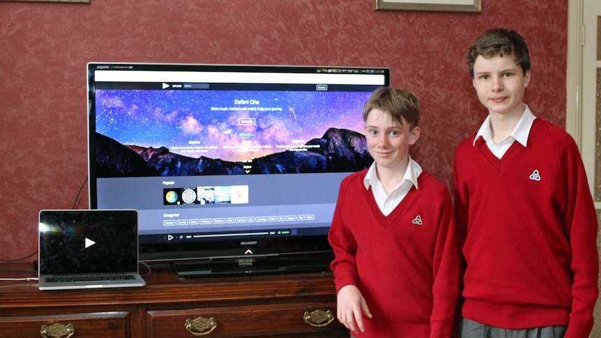Two 14 year-old-boys stand side by side next to a tv screen with website displayed