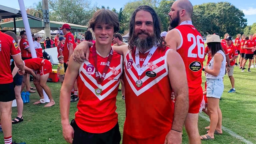 A son and father in arms wearing a red and white football uniform and a winning medal around their necks.