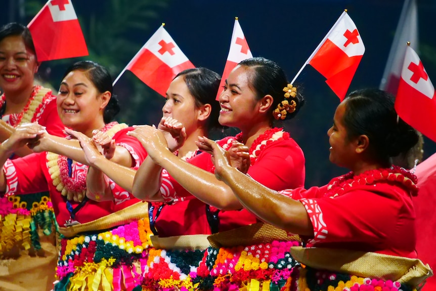 Women wearing red dance while Tongan flags flutter above.