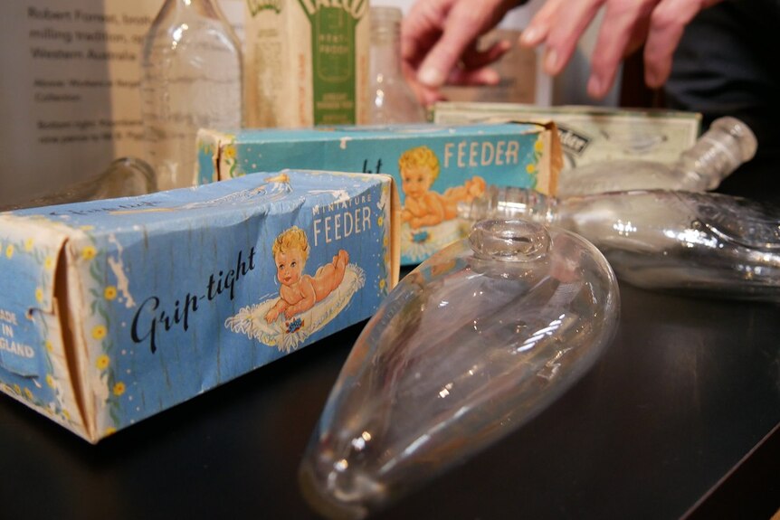 Several old baby bottles with their box on a table with a man's hands in the background.