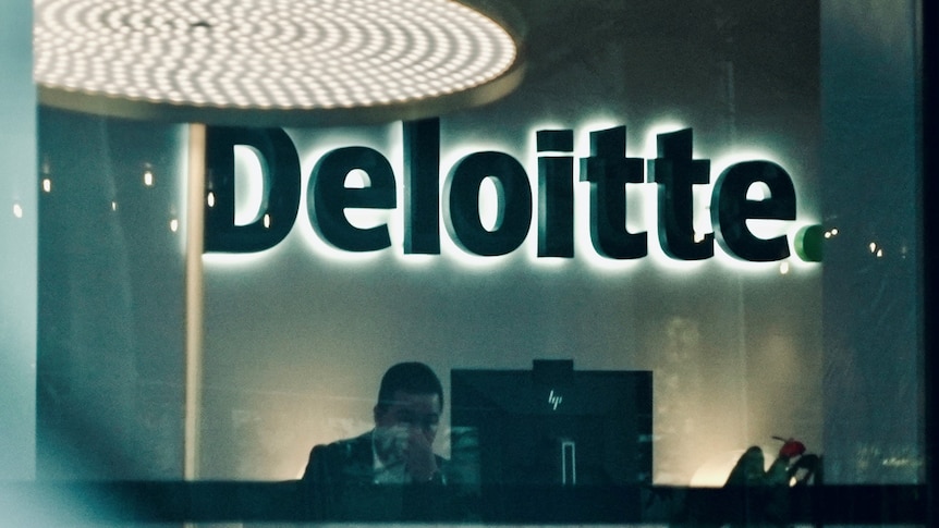 An illuminated Deloitte logo sits above a desk with a worker sitting behind a computer screen.