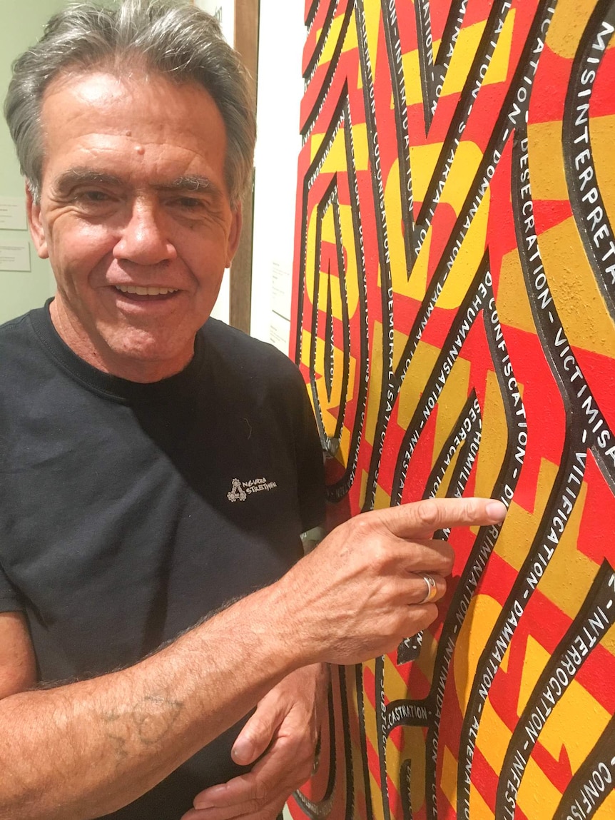 Grey haired man wearing a black T-shirt standing alongside his red, yellow, black and white artwork of snaking lines over words