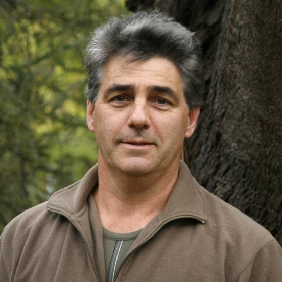 Man in brown jumper stands against tree and smiles at camera