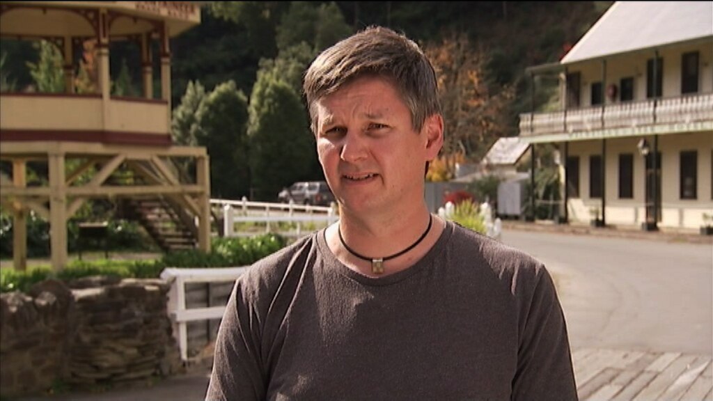 Man with brown long sleeve shirt and necklace standing in front of old buildings in Walhalla 