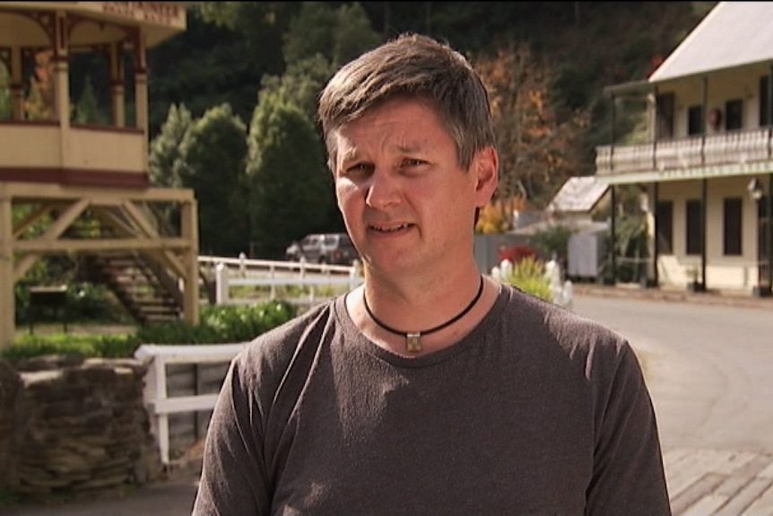 Man with brown long sleeve shirt and necklace standing in front of old buildings in Walhalla 